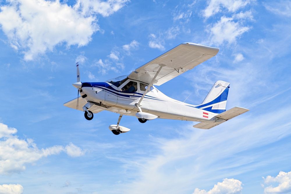 White and blue single-engine airplane flying in a blue sky with scattered clouds.