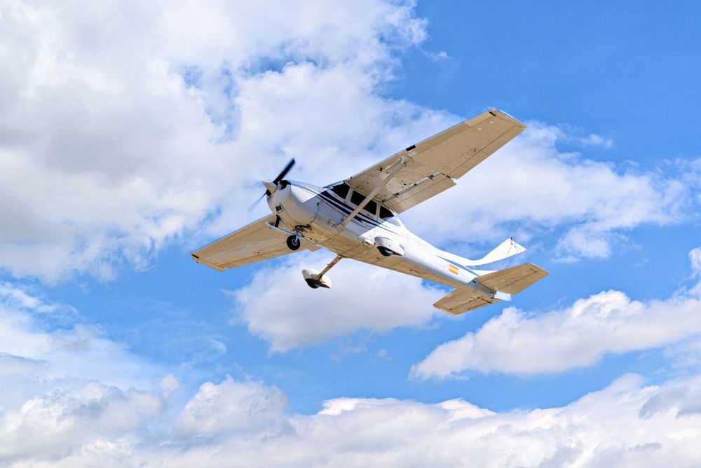 A white light aircraft flying through a blue sky with scattered white clouds.