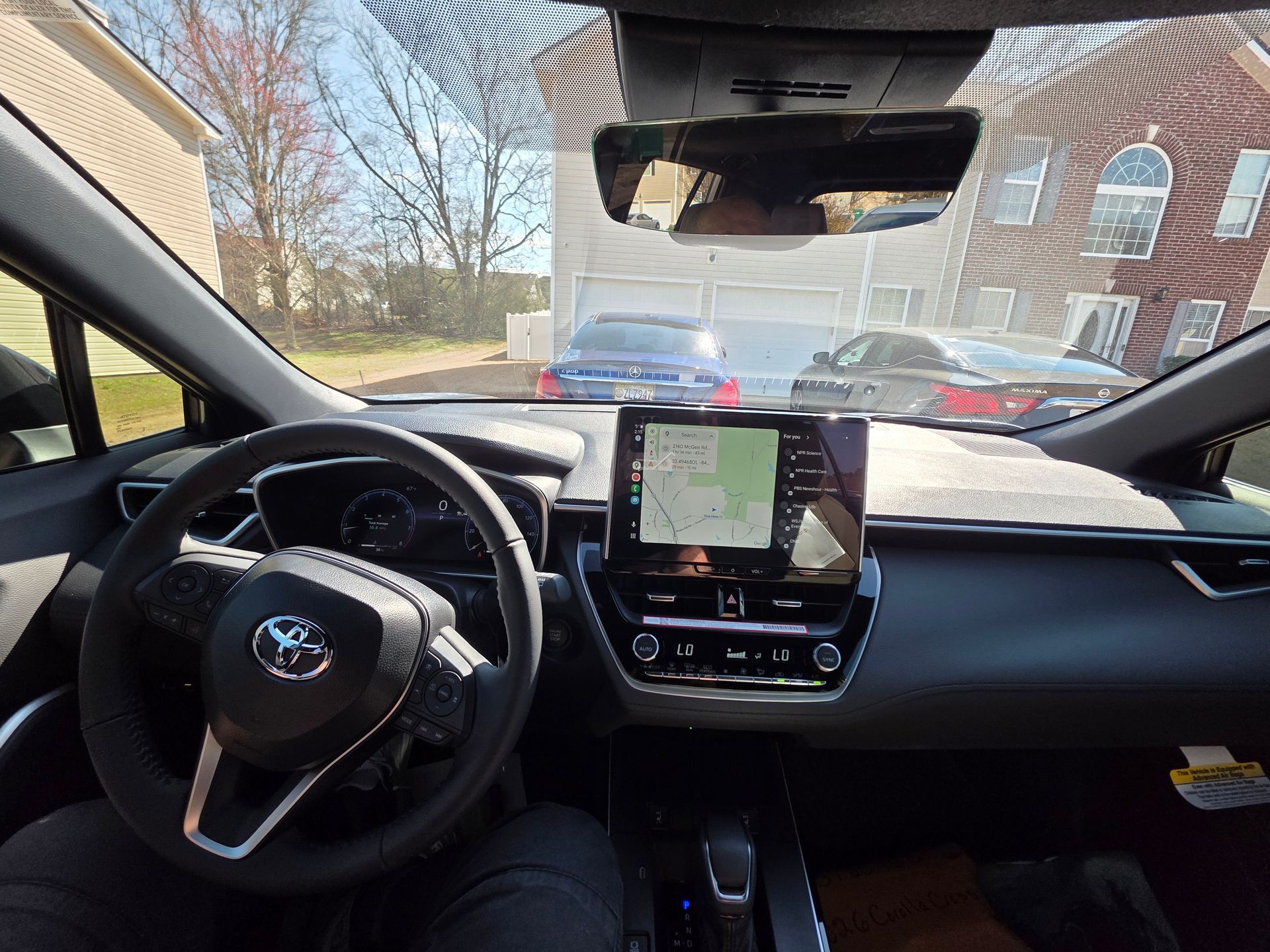 Driver's view from inside a Toyota, showing the steering wheel, dashboard, infotainment screen, and a sunny street ahead.
