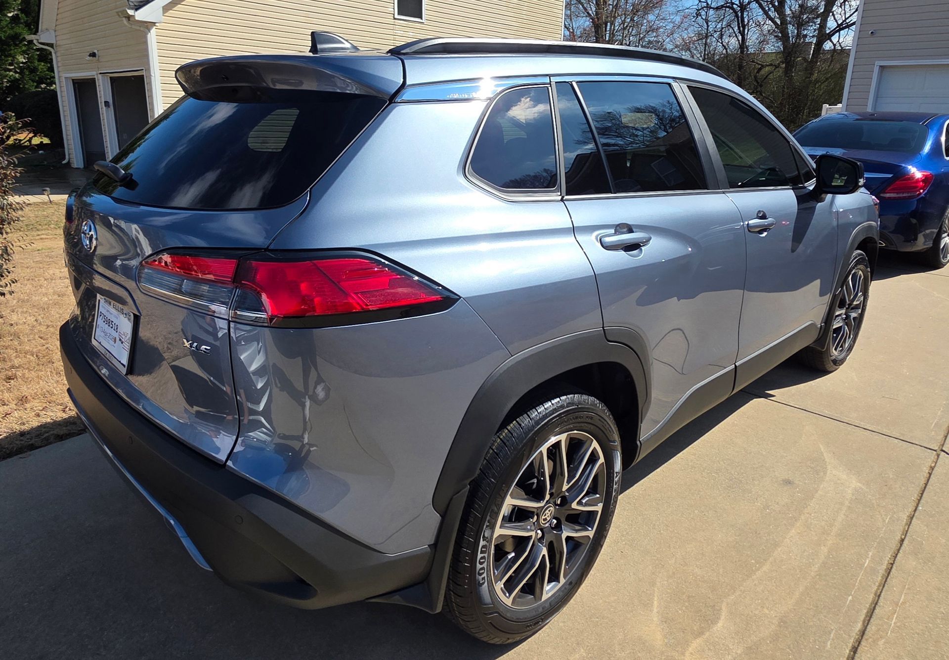 A metallic light blue Toyota Corolla Cross SUV parked on a concrete driveway in a suburban residential setting.