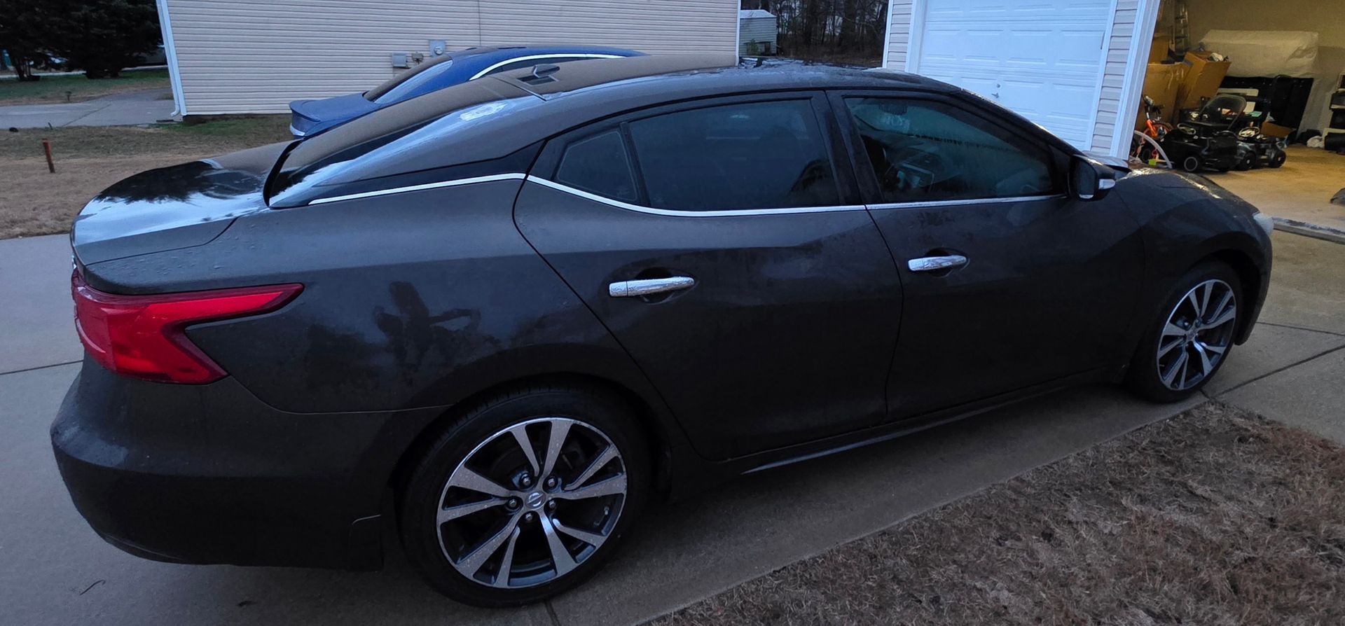 A charcoal gray Nissan Maxima parked on a driveway next to a residential garage.