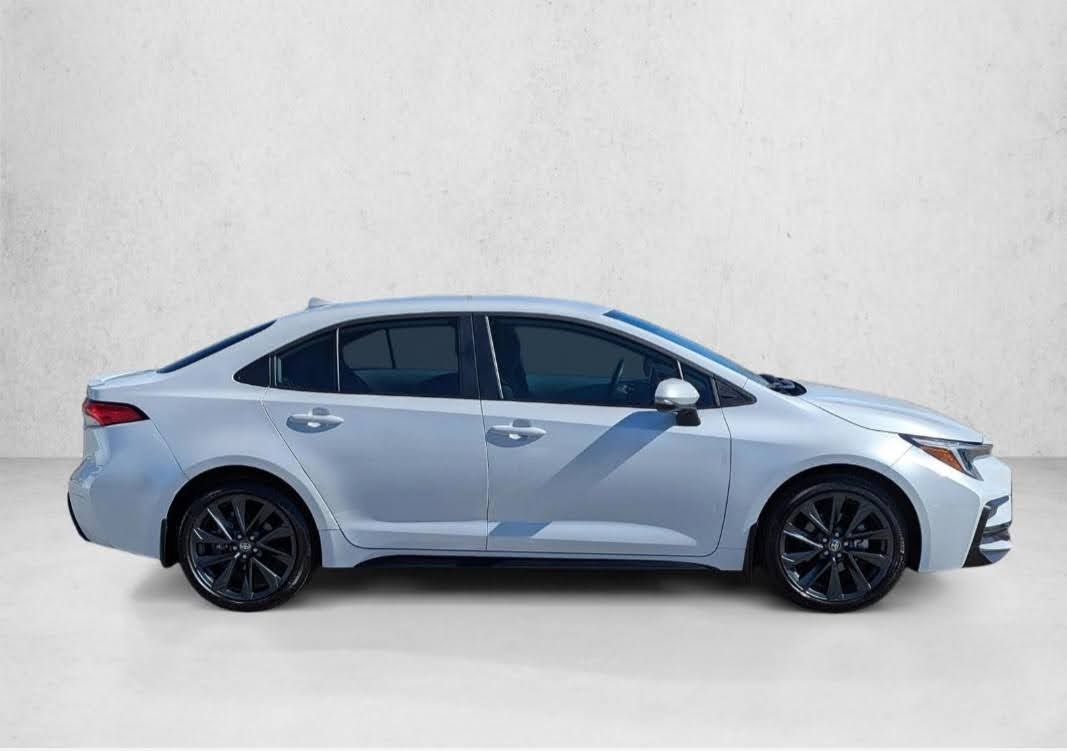 A silver Toyota Corolla sedan seen from the side profile, featuring dark wheels and tinted windows against a white wall.