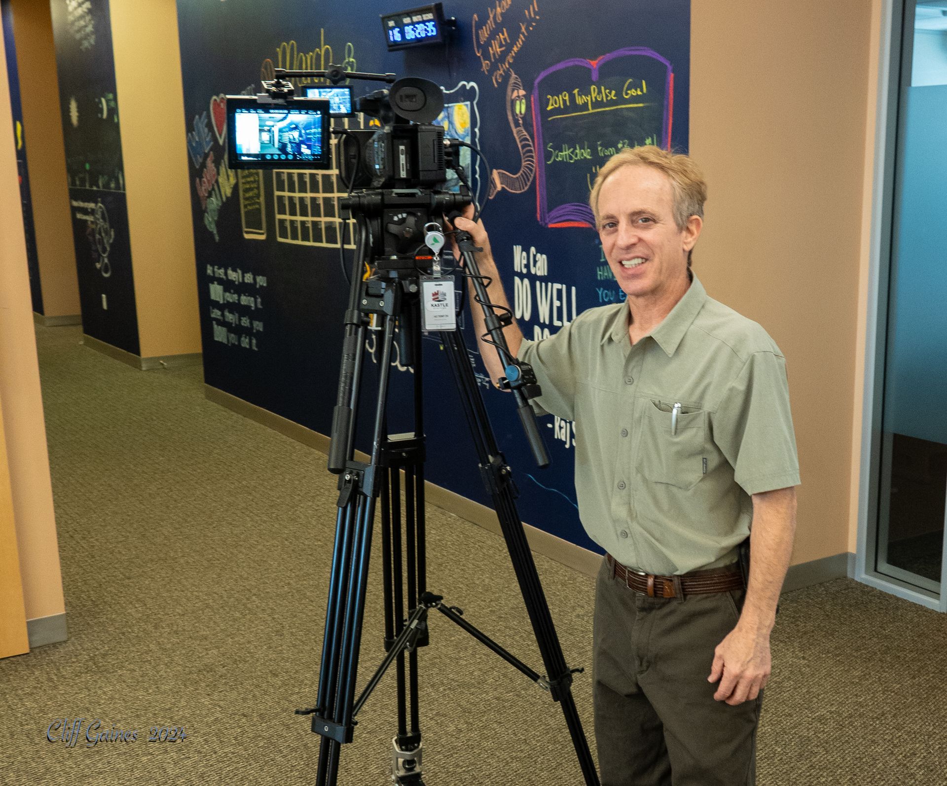 Cliff stands in front of a camera with a sign on the wall that says we can