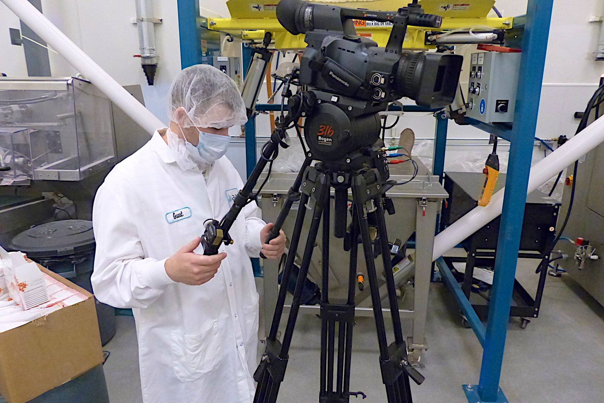 A man wearing a mask stands in front of a camera on a tripod