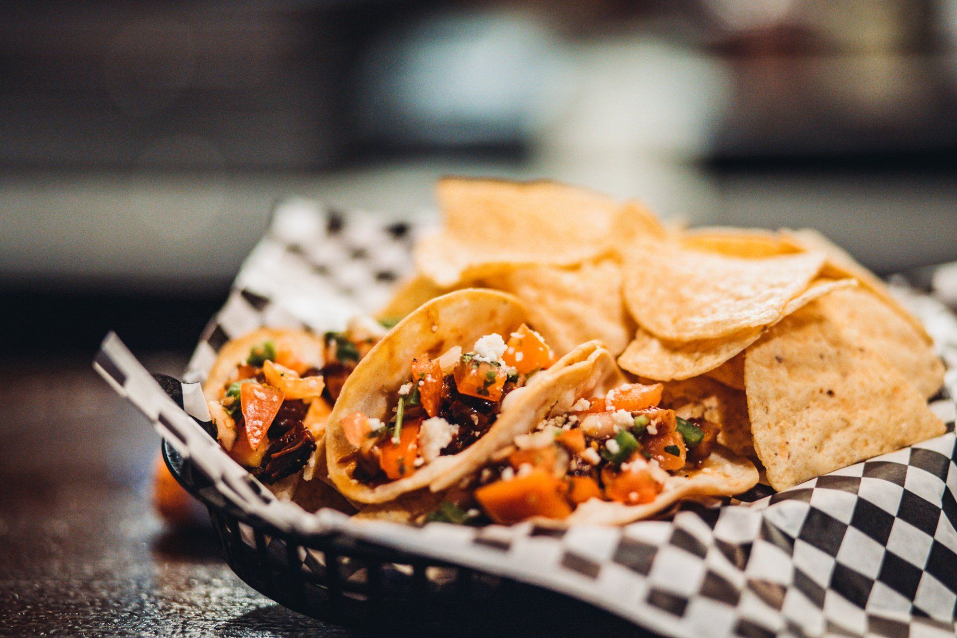 A basket of tacos and chips on a checkered paper on a table.