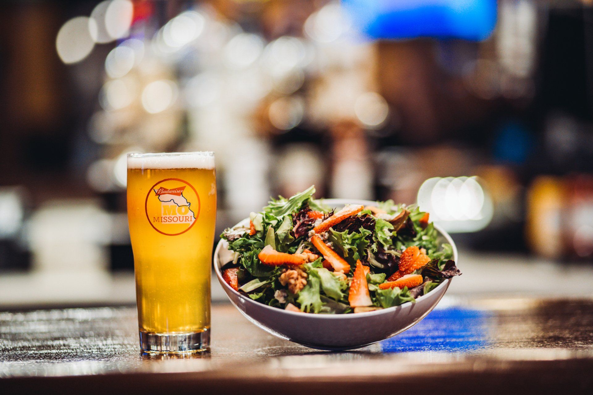 A bowl of salad and a glass of beer on a bar.