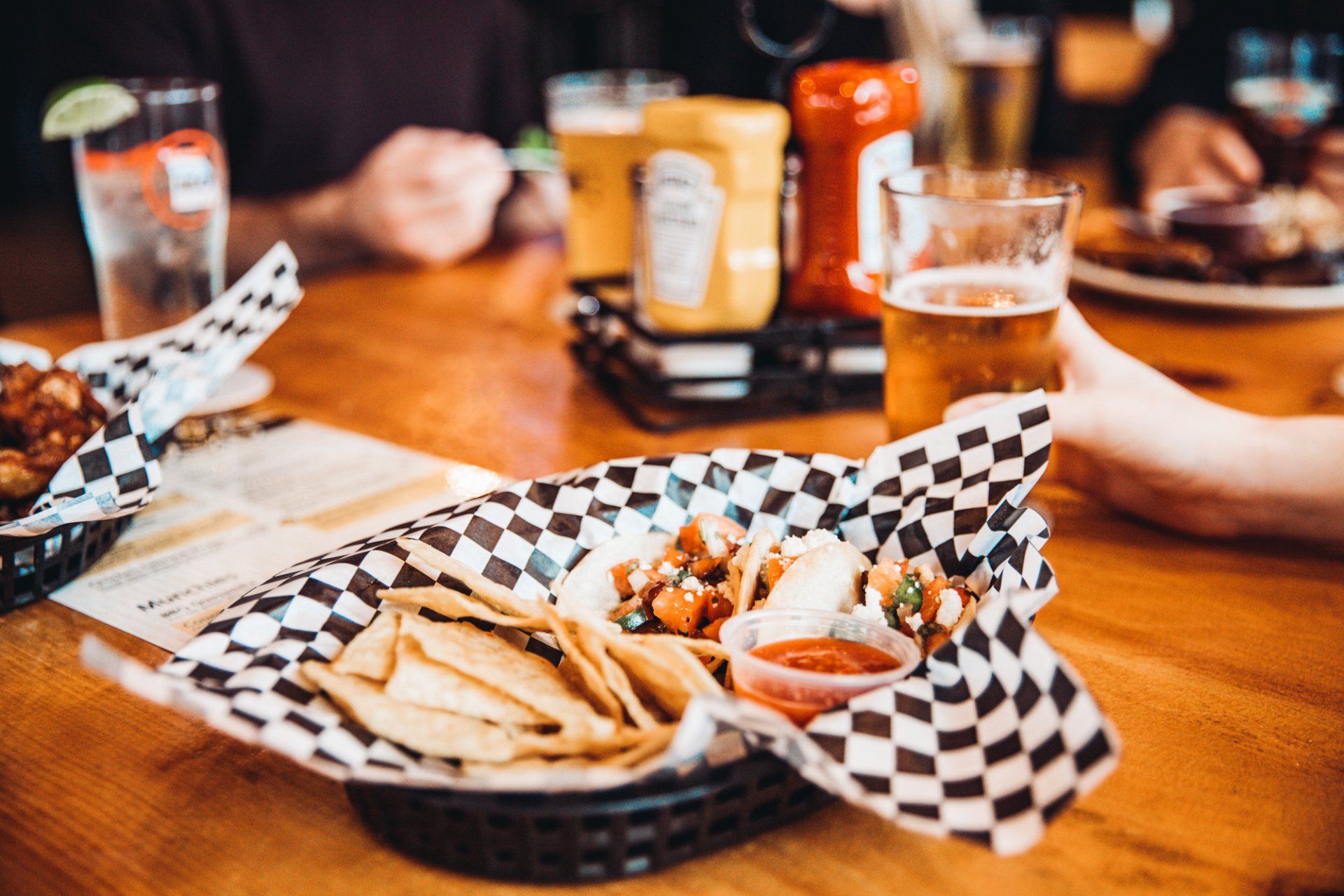 A person is holding a glass of beer and a basket of food on a table.