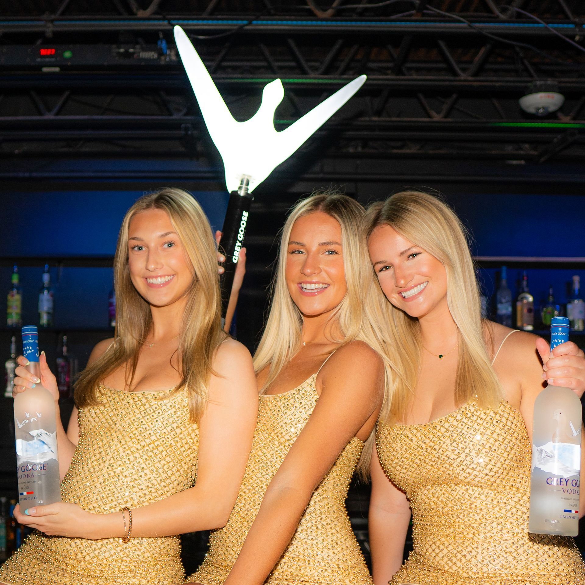 Three women in gold dresses are holding bottles of vodka