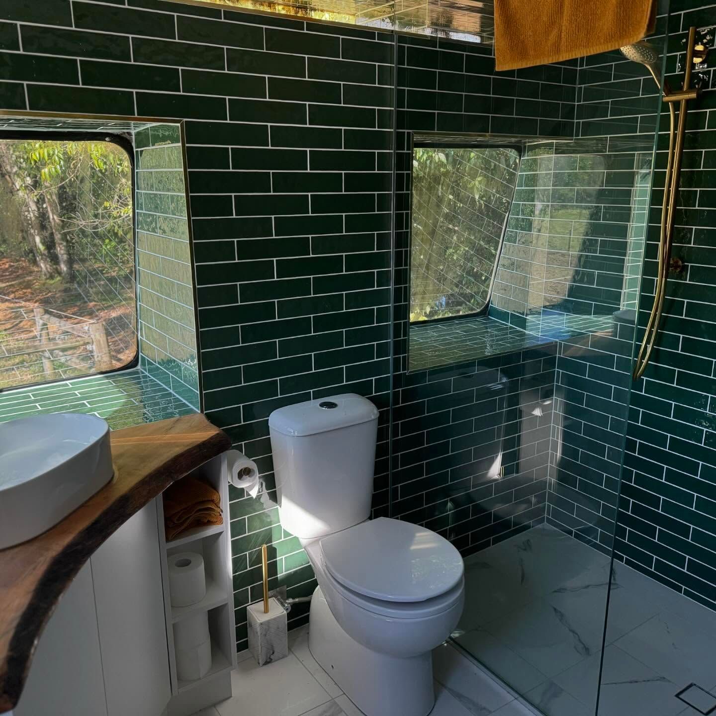 Green-tiled bathroom in a bus conversion. Features a toilet, sink, shower, and two windows overlooking a forest.