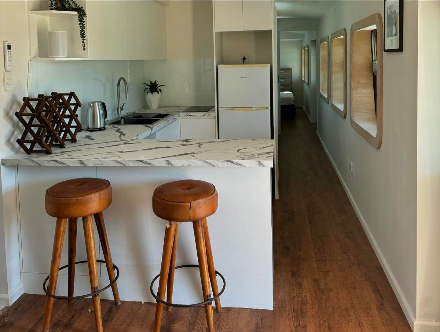A small kitchen with a marble countertop, two wooden stools, and a refrigerator. A hallway extends from the kitchen with framed artwork.