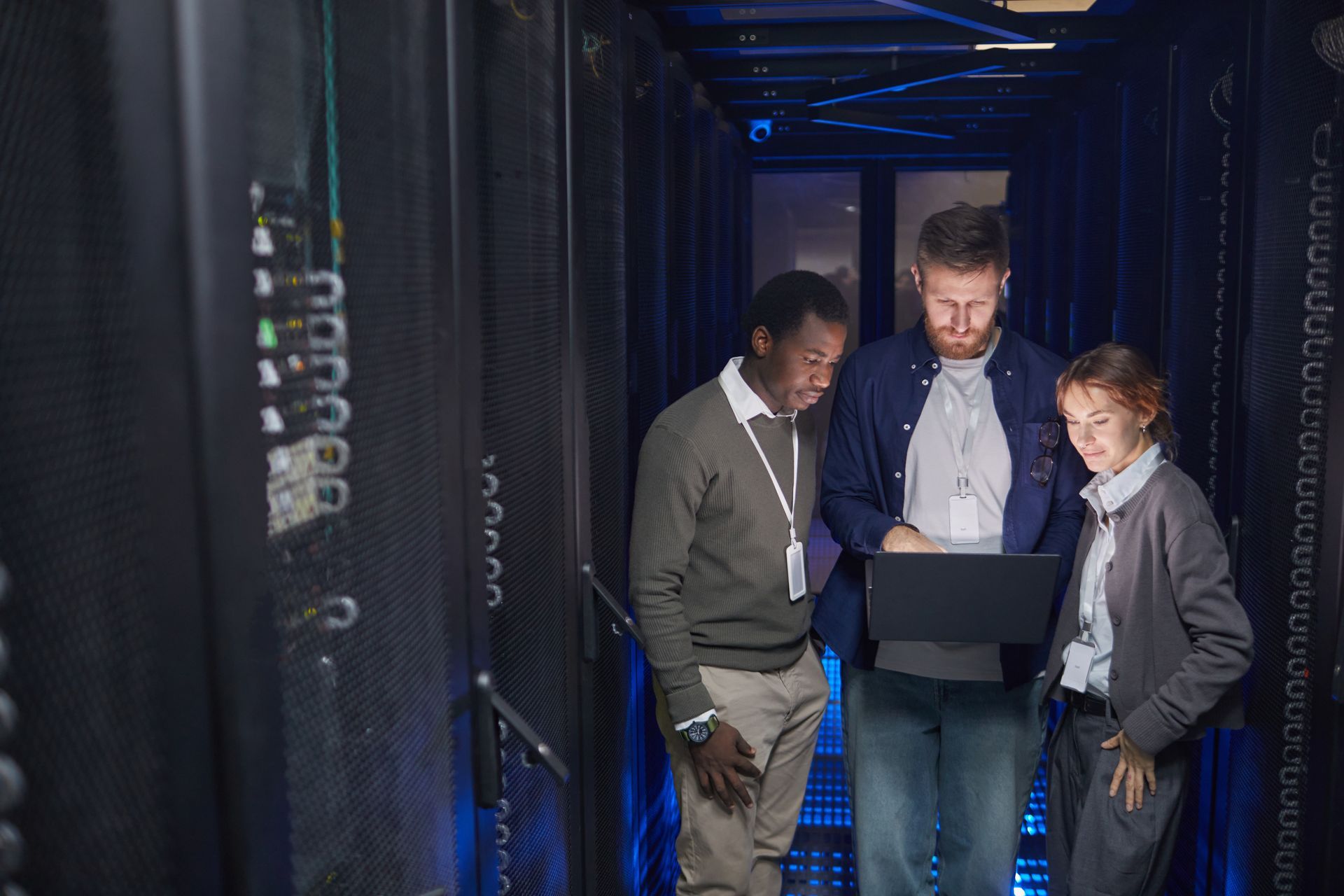 Three people looking at laptop in server room; dark, with blue lights.