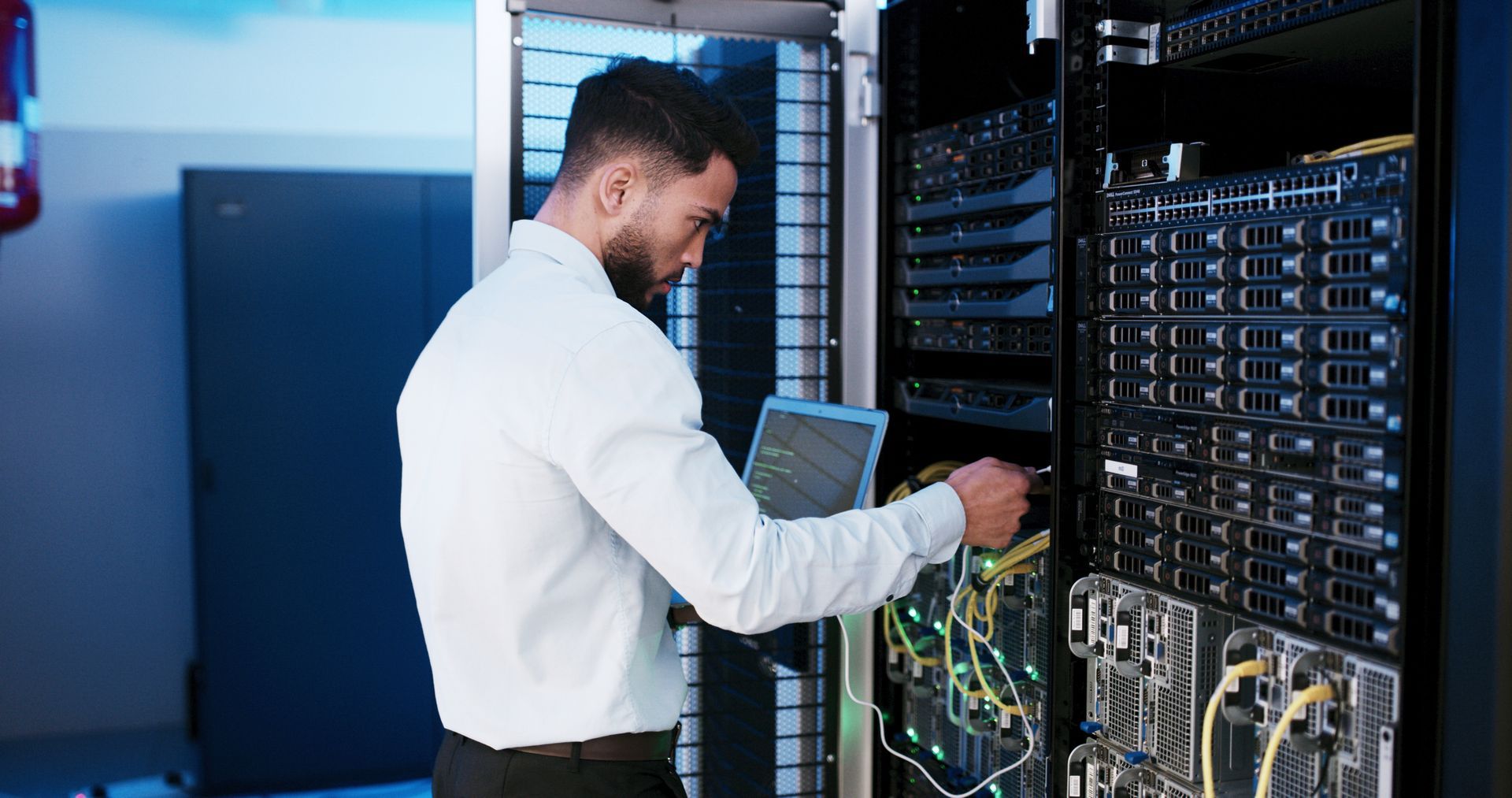 Man working on server rack in a data center, holding a tablet and connecting cables.