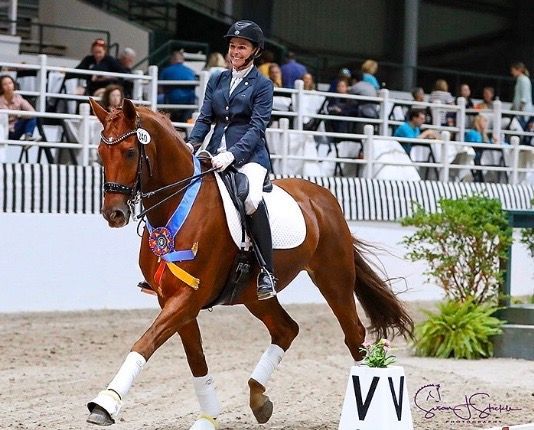 A woman is riding a brown horse with a blue ribbon around its neck