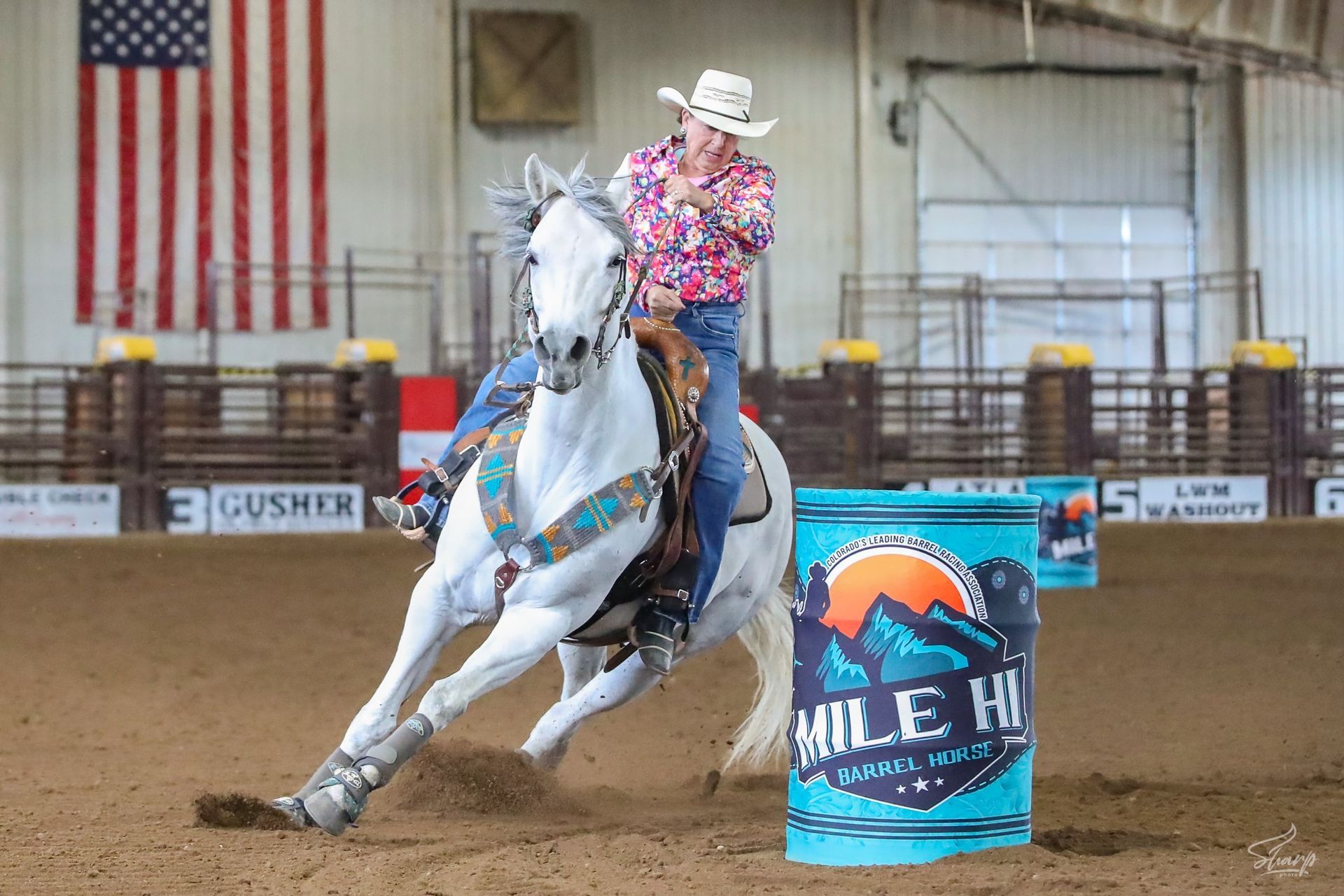 A woman is riding a white horse around a blue barrel.