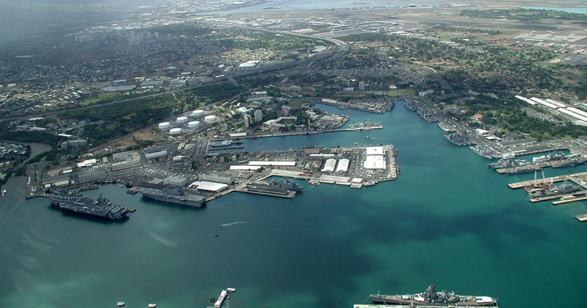 Aerial view of a harbor with numerous ships and docks, against a backdrop of a city and blue ocean.