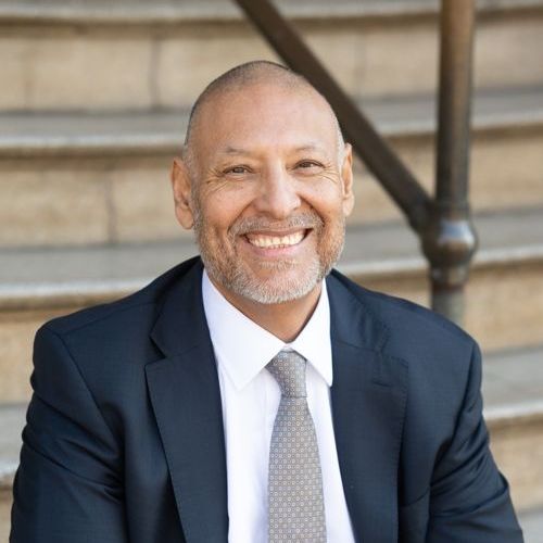 Smiling man in a suit and tie sits on steps, looking at the camera.