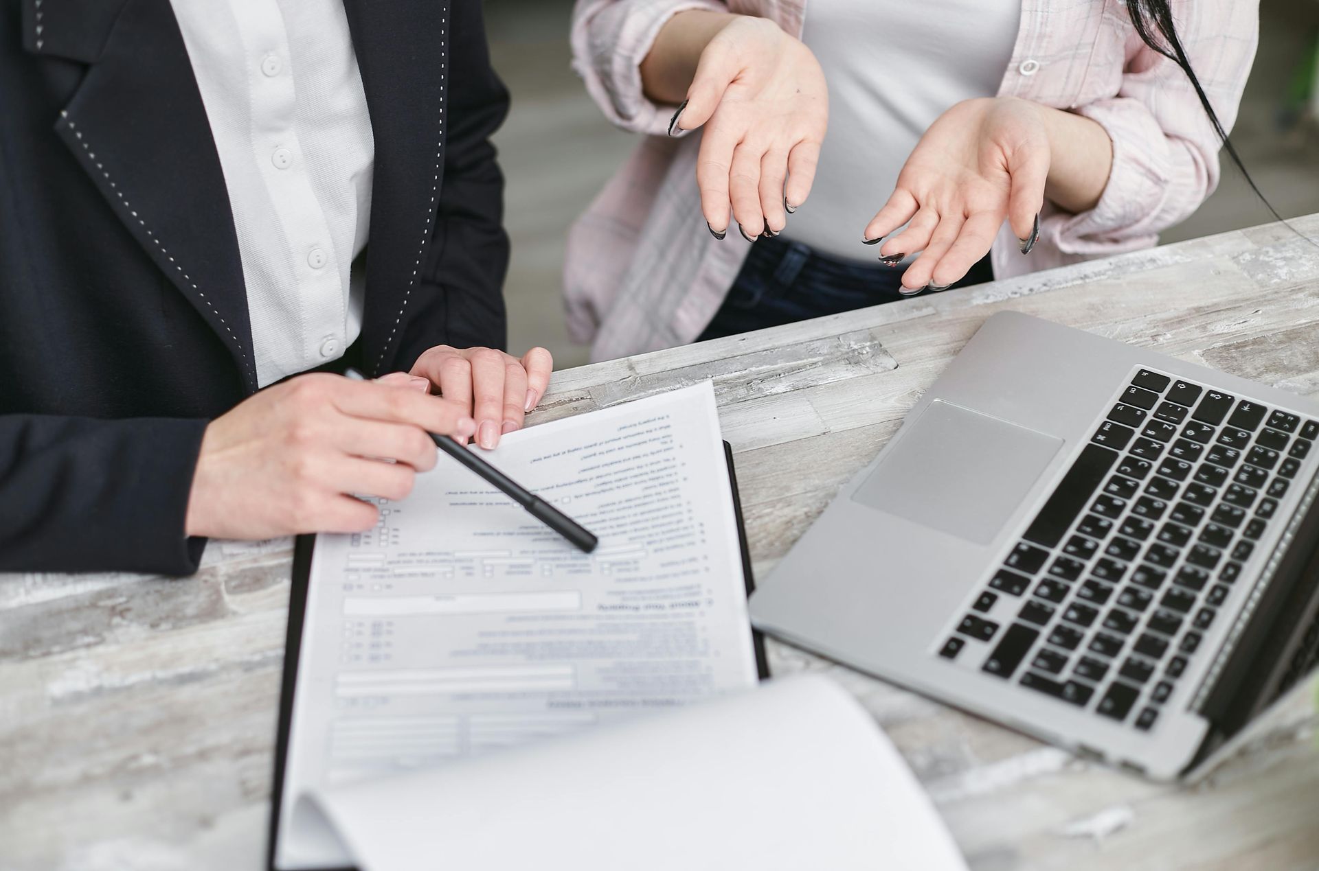 Two women at a table, one pointing at a form while the other gestures with hands. Laptop and papers present.