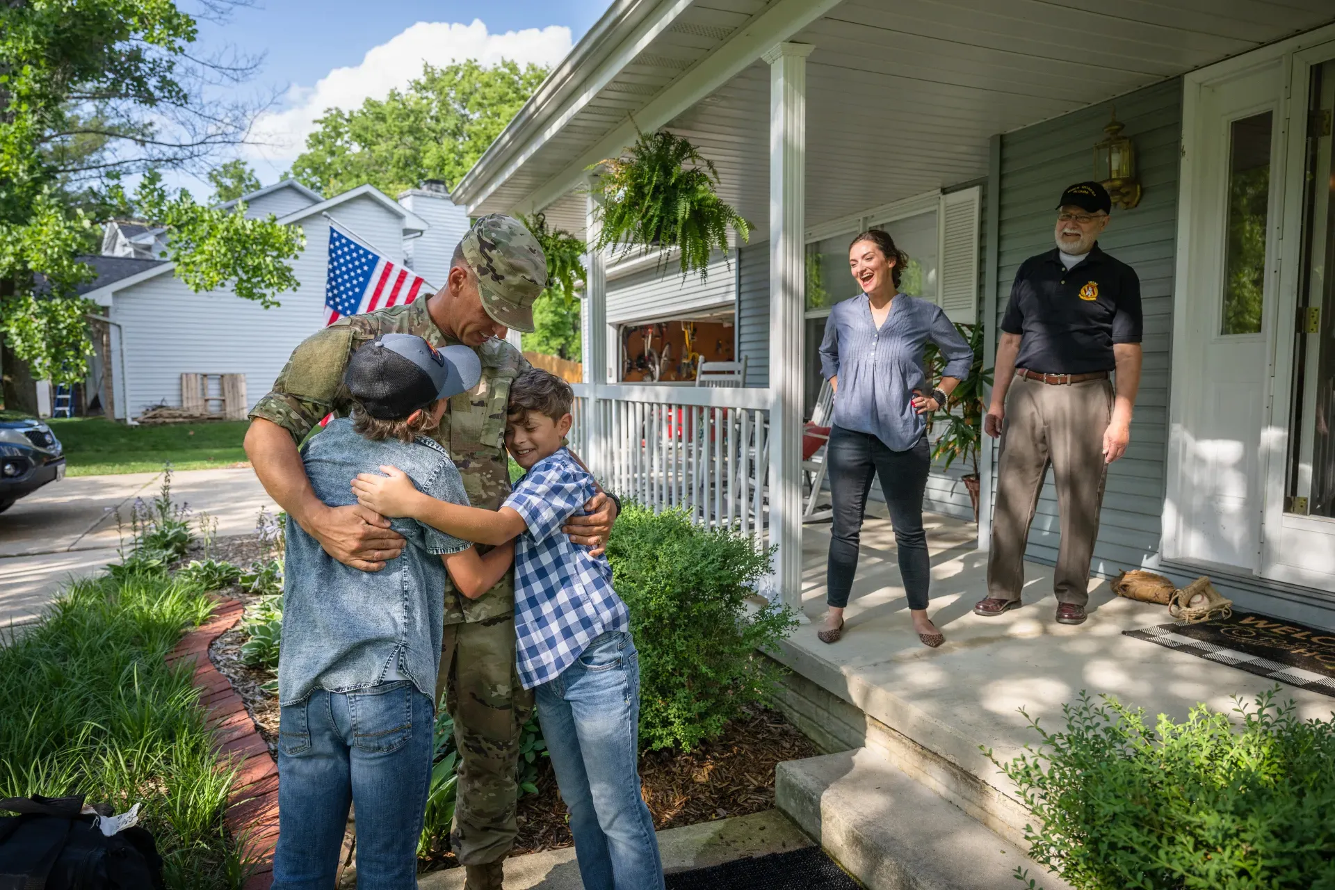 Soldier hugging two children outside a house; mother and another man watch on the porch. American flag visible.