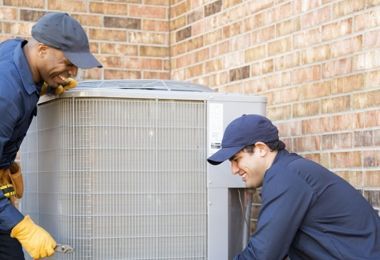 Two men are working on an air conditioner outside of a brick building.