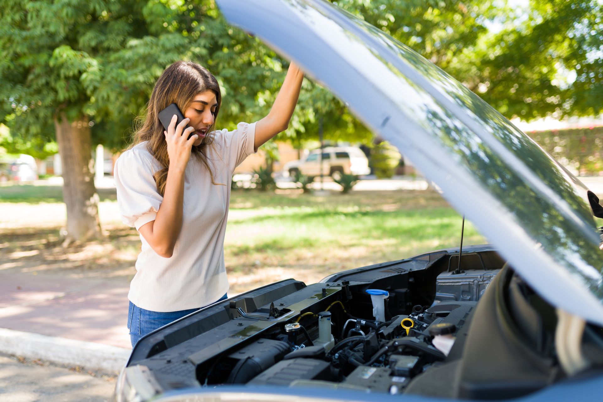 A woman is talking on a cell phone while looking under the hood of her broken down car.