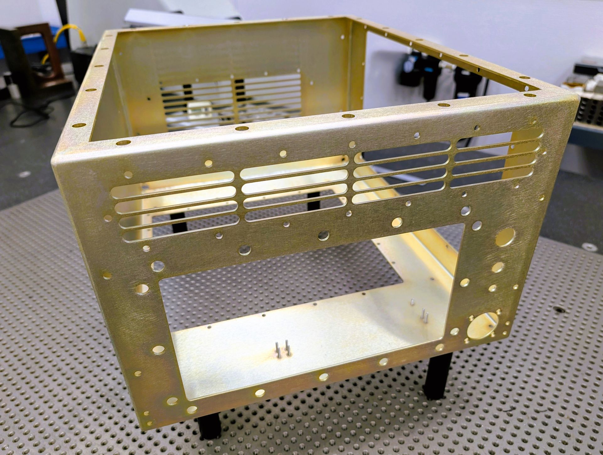 Gold metal electronic equipment enclosure with ventilation slots and mounting holes, on black legs.
