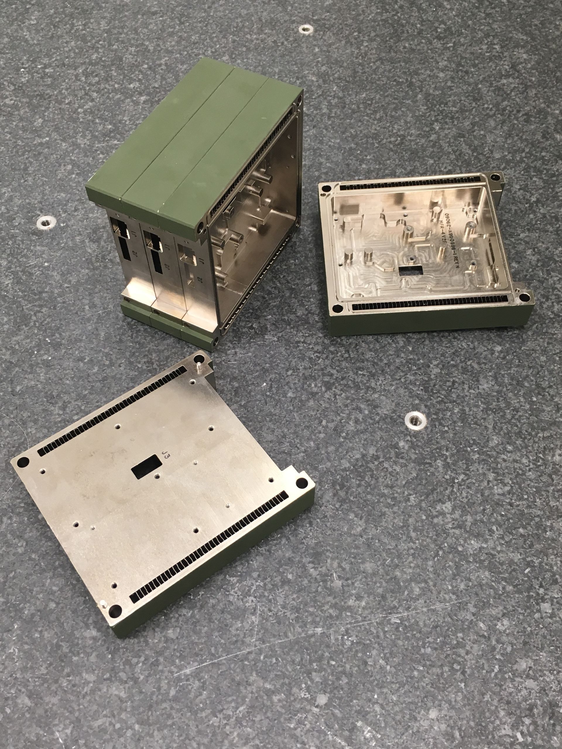 Three open, rectangular, metal electronic device components with olive drab painted edges on a grey surface.