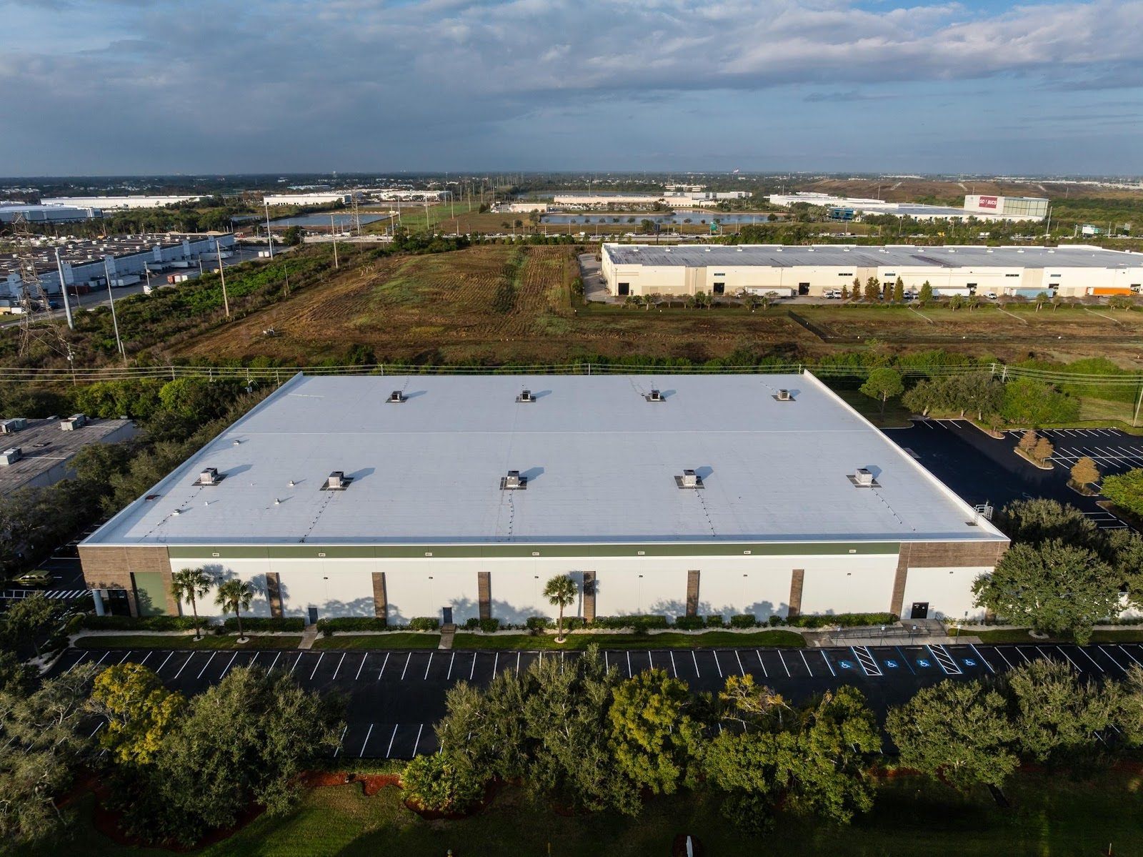 Aerial view of a large warehouse with a flat roof, surrounded by trees, parking lot, and other buildings.