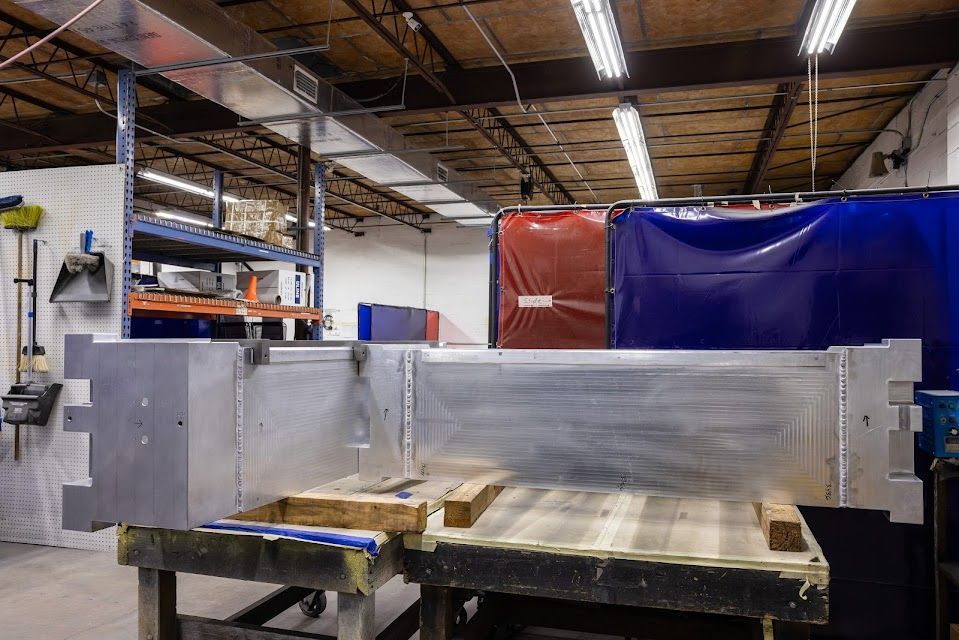 Metal fabrication workshop: Large aluminum structure on work table, welding screens in background, shelving unit.