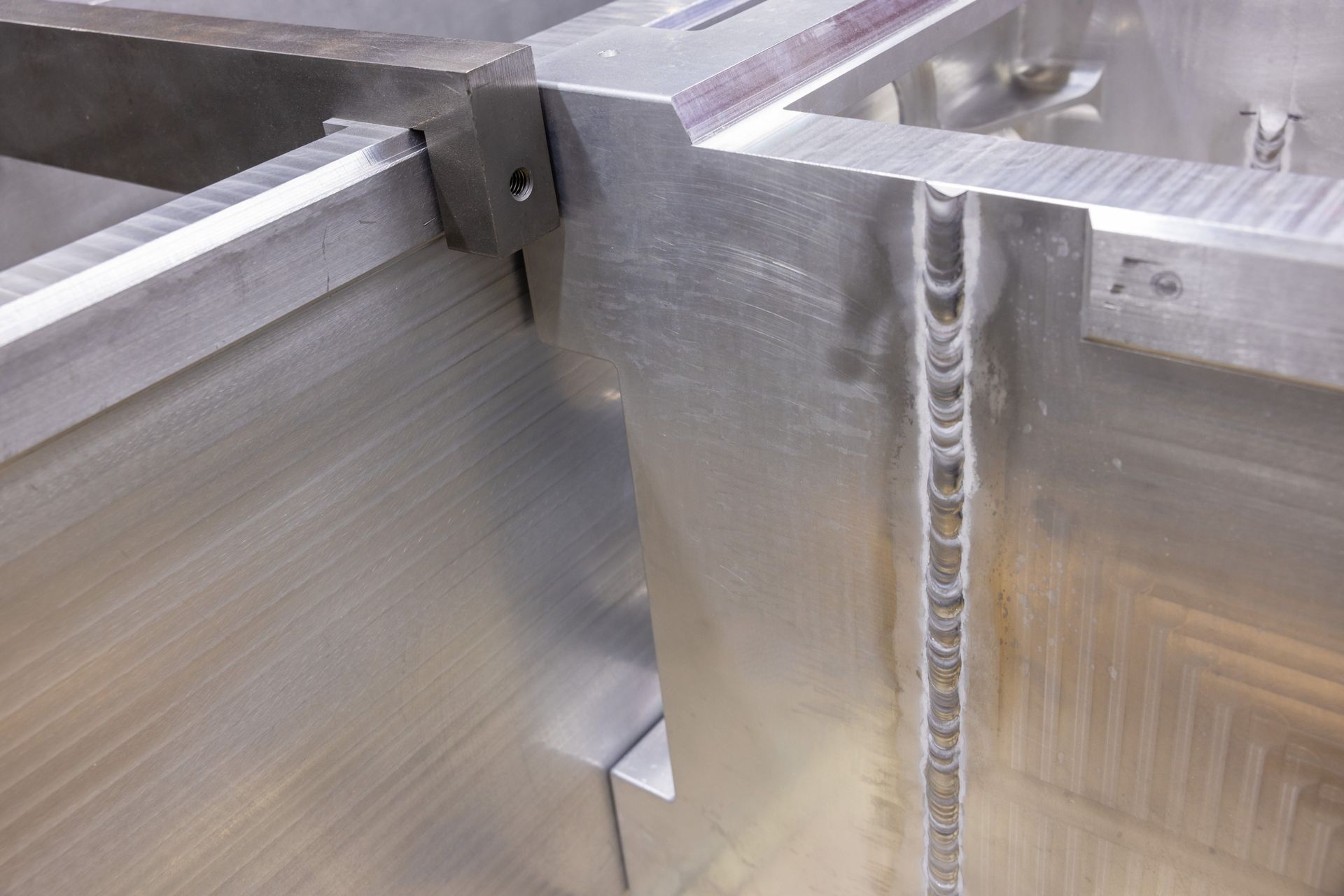 Close-up of welded aluminum frame corner. Silver metal with visible weld marks and machining lines.