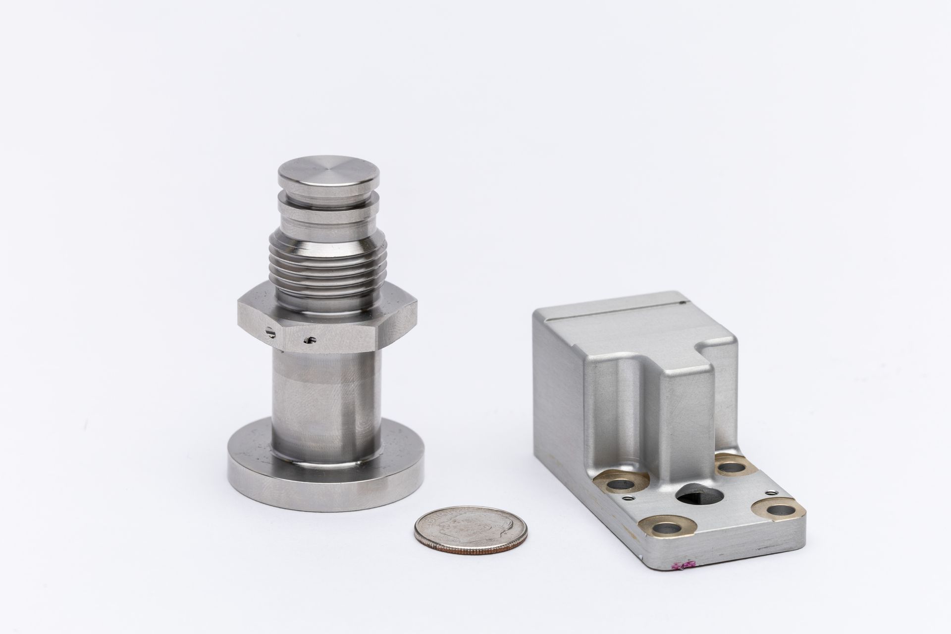 Silver mechanical components next to a coin, on white. One component is a bolt, the other is a small block.