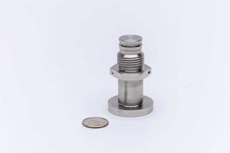 Silver metal bolt next to a US quarter coin, with a threaded section and circular base.