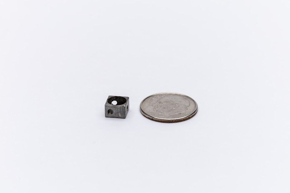 Small, square, metal object next to a coin on a white surface.