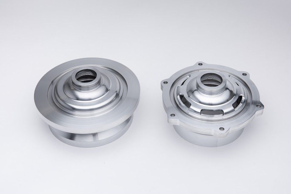 Two shiny, metallic, circular mechanical parts on a white background. One is a solid disc with a central hex. The other has exposed interior mechanics.