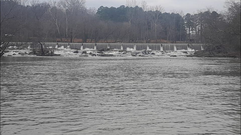 A wide, choppy river flows towards a small dam. Trees line the background under a cloudy sky.