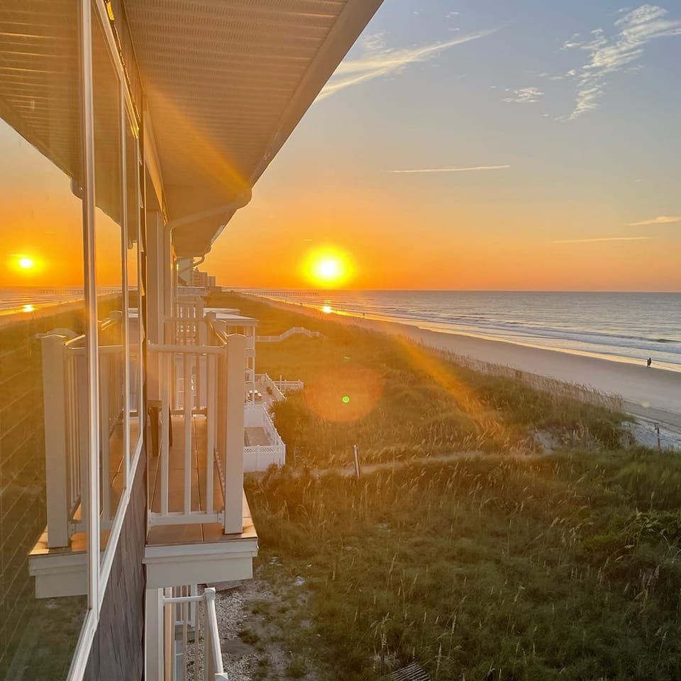 Sunset over the ocean, viewed from a balcony. Orange sky, beach, waves, and a person walking.