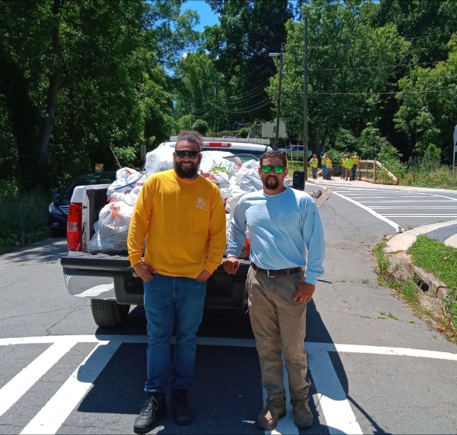 Two men stand by a truck filled with trash bags on a road. One wears yellow, the other blue.