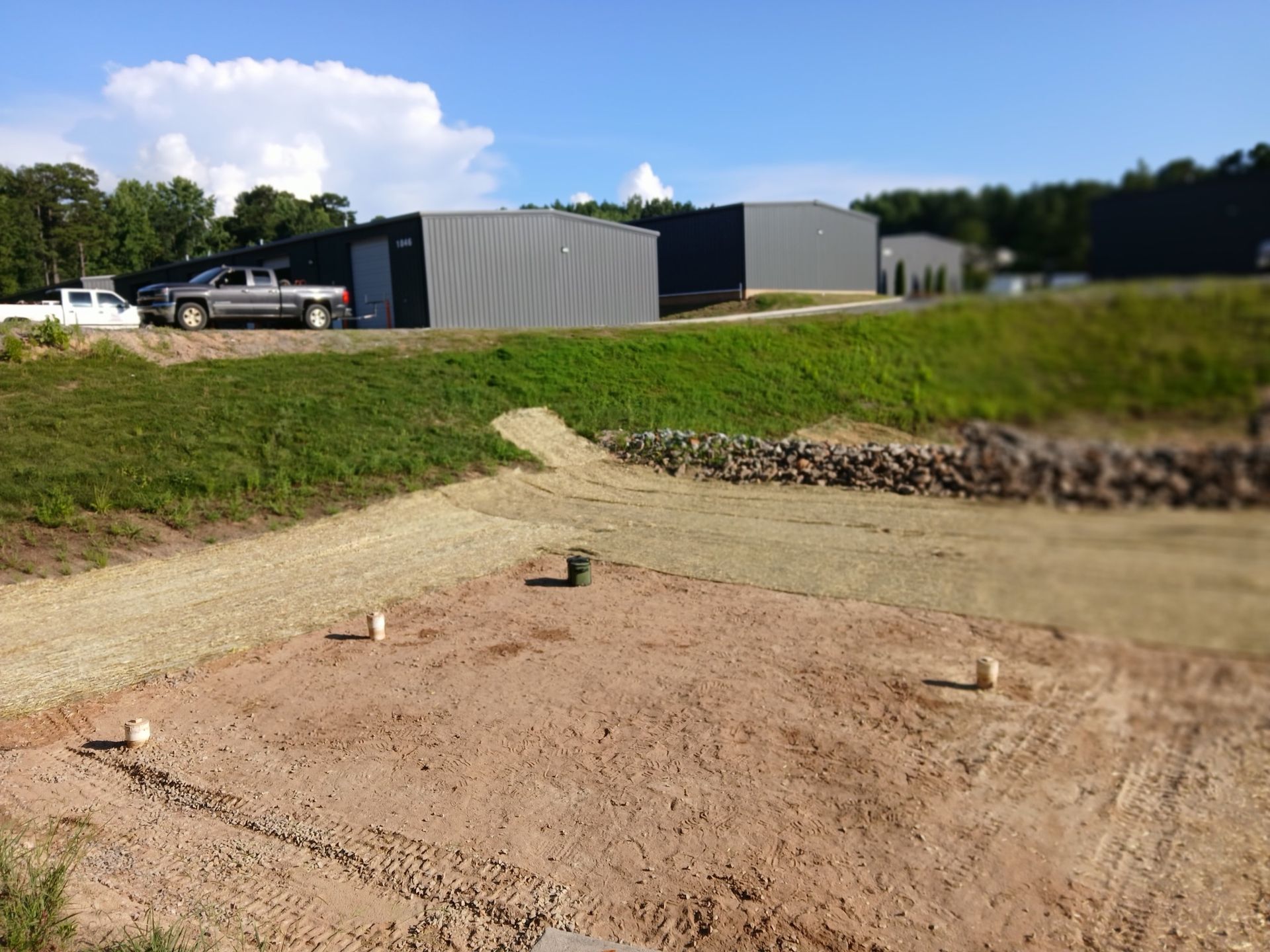 Dirt patch with gravel path and three white posts, leading toward gray metal buildings and a pickup truck.
