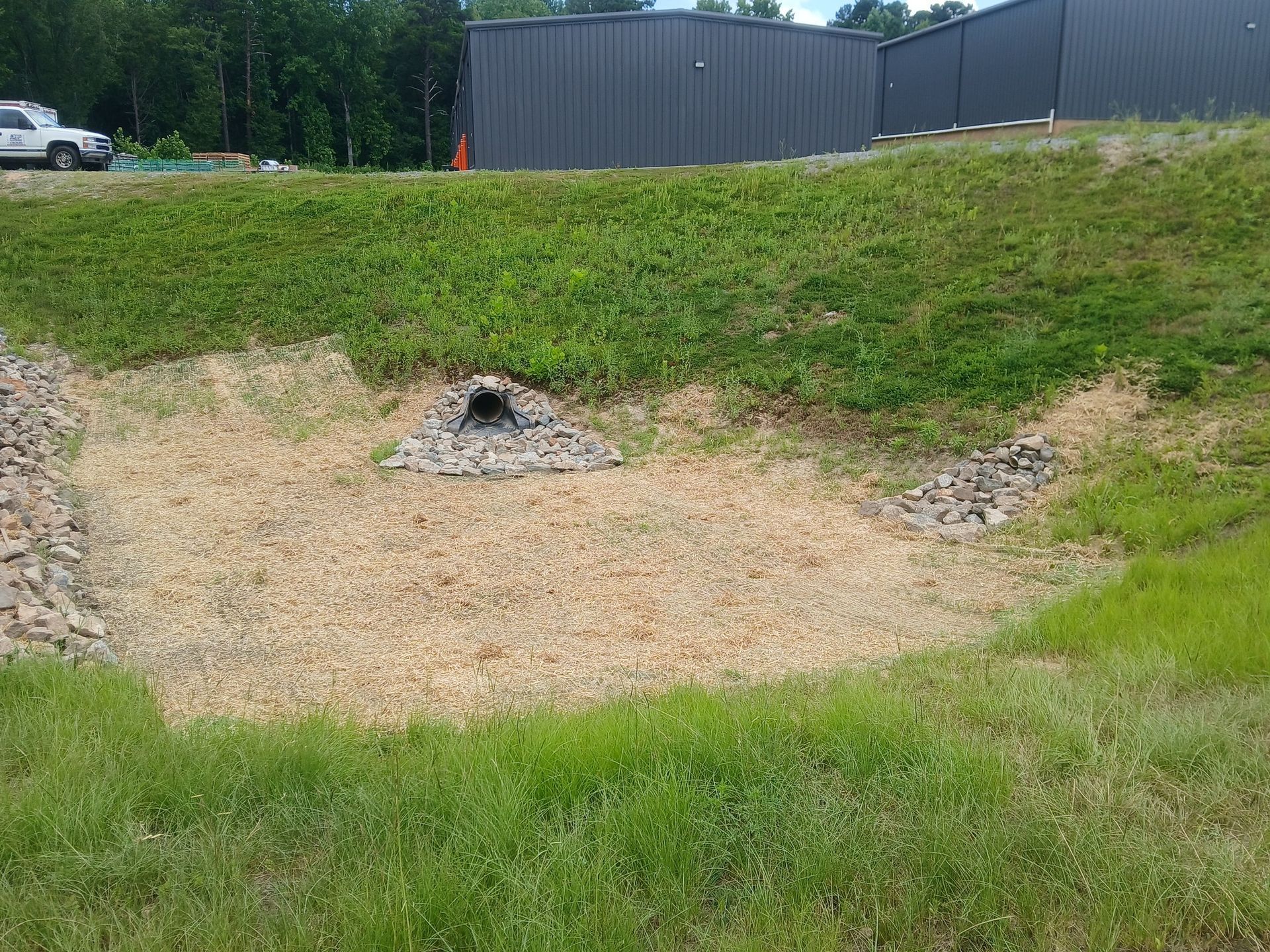 A drainage basin with a culvert and gravel, surrounded by grass. A building is in the background.