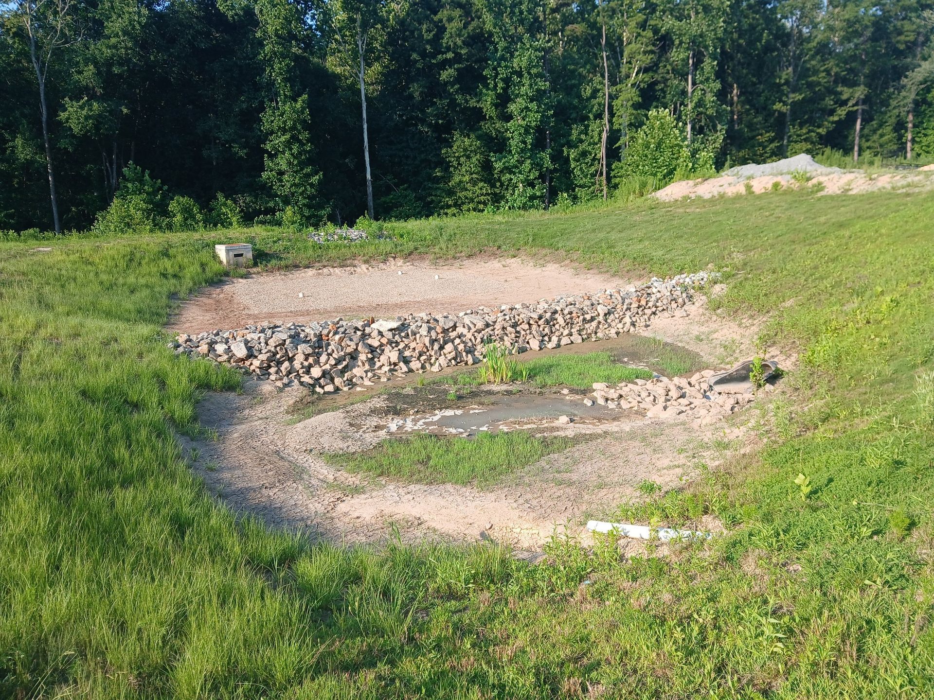 A circular dry pond surrounded by grass and trees, with stone barriers and a small water area.