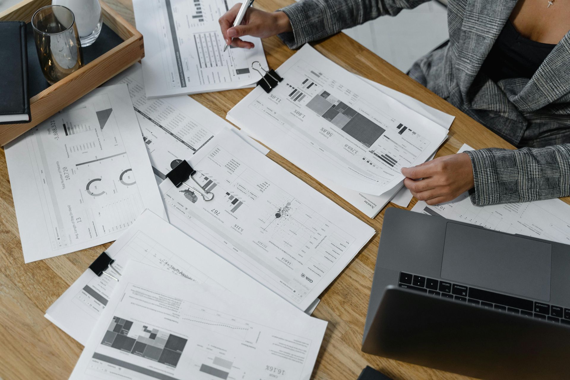 Person in blazer reviews documents with graphs and a laptop on a desk.