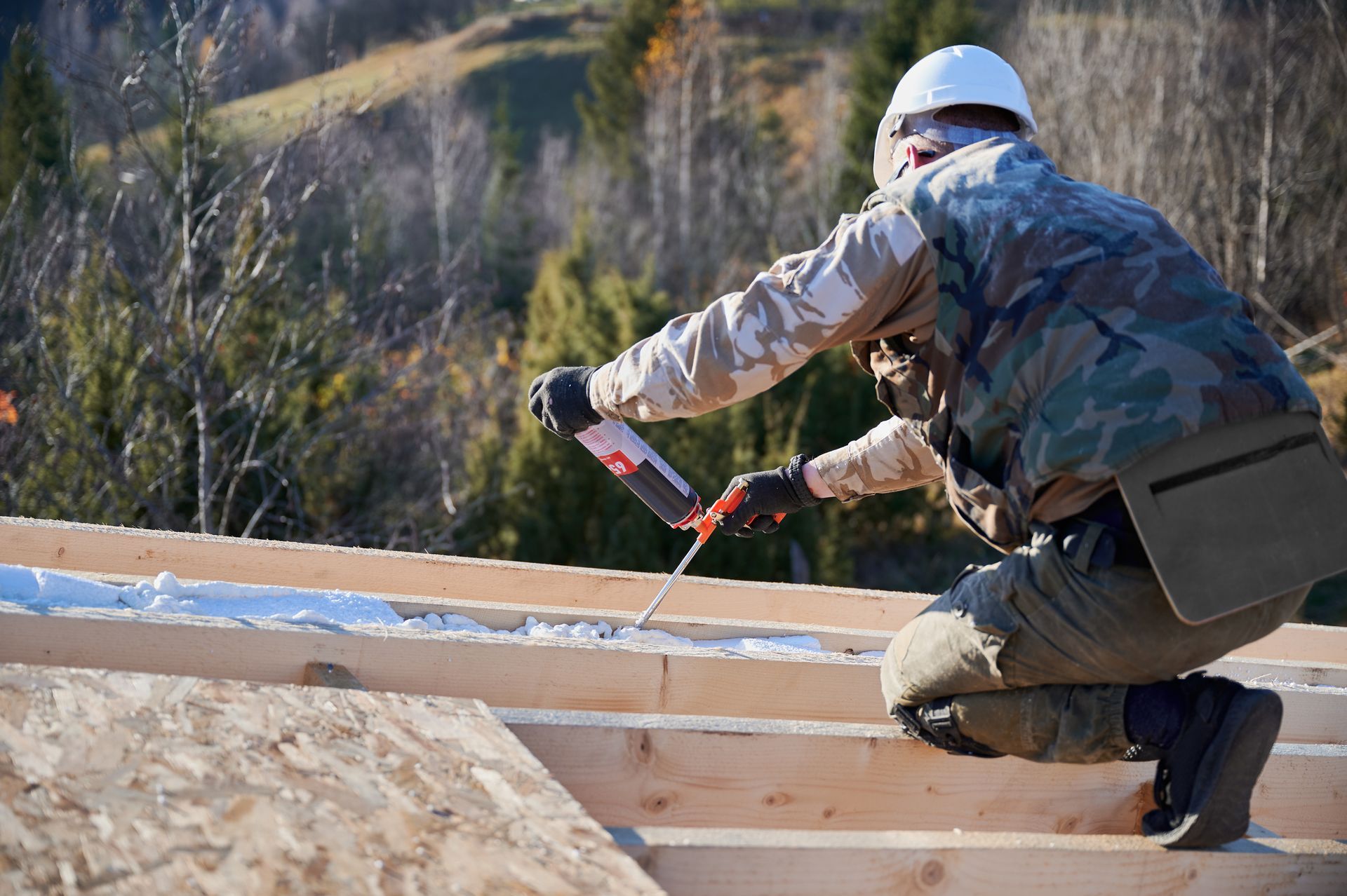 Worker in helmet sealing wooden beams at rural construction site.