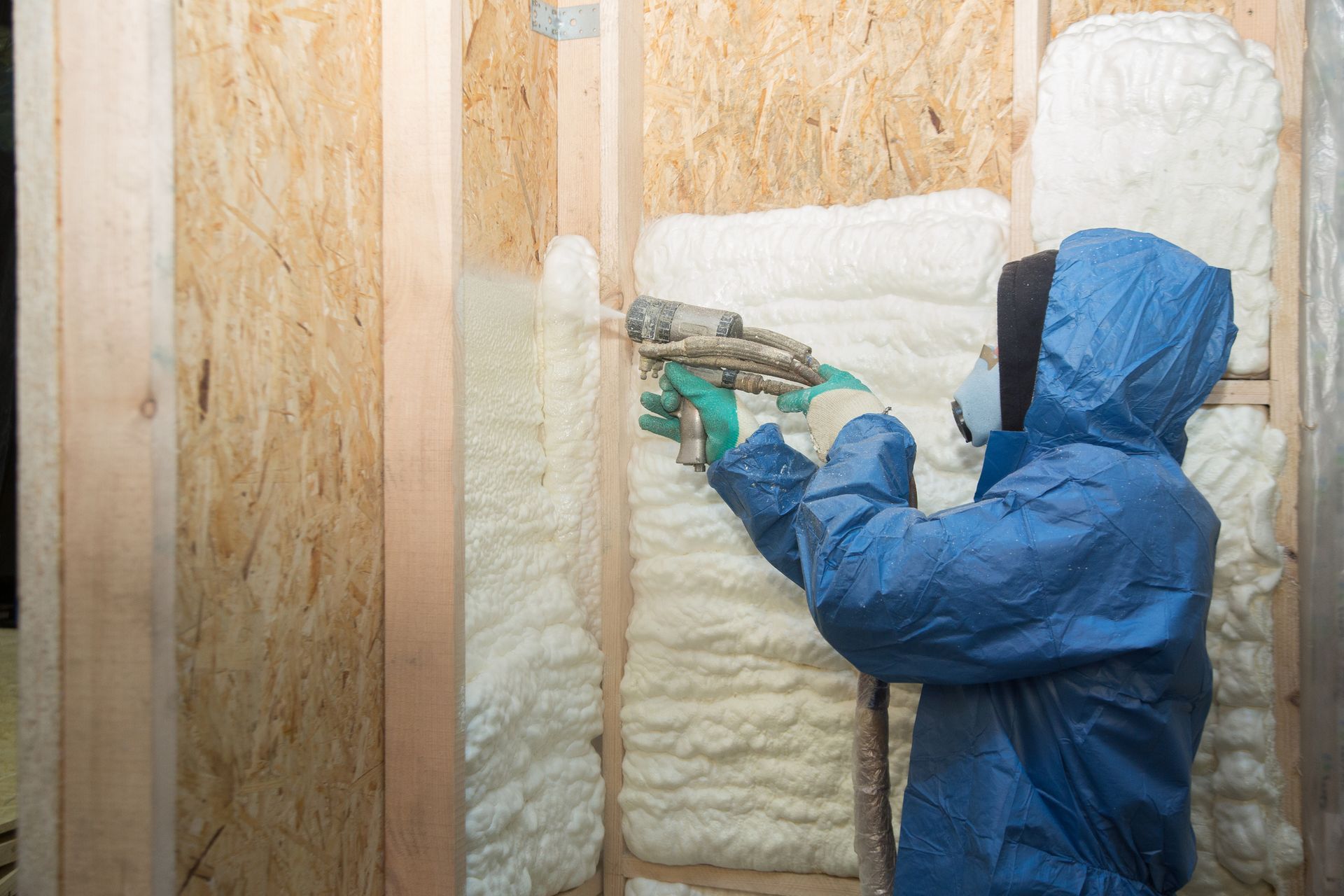 A person spraying foam insulation on a wooden wall frame.