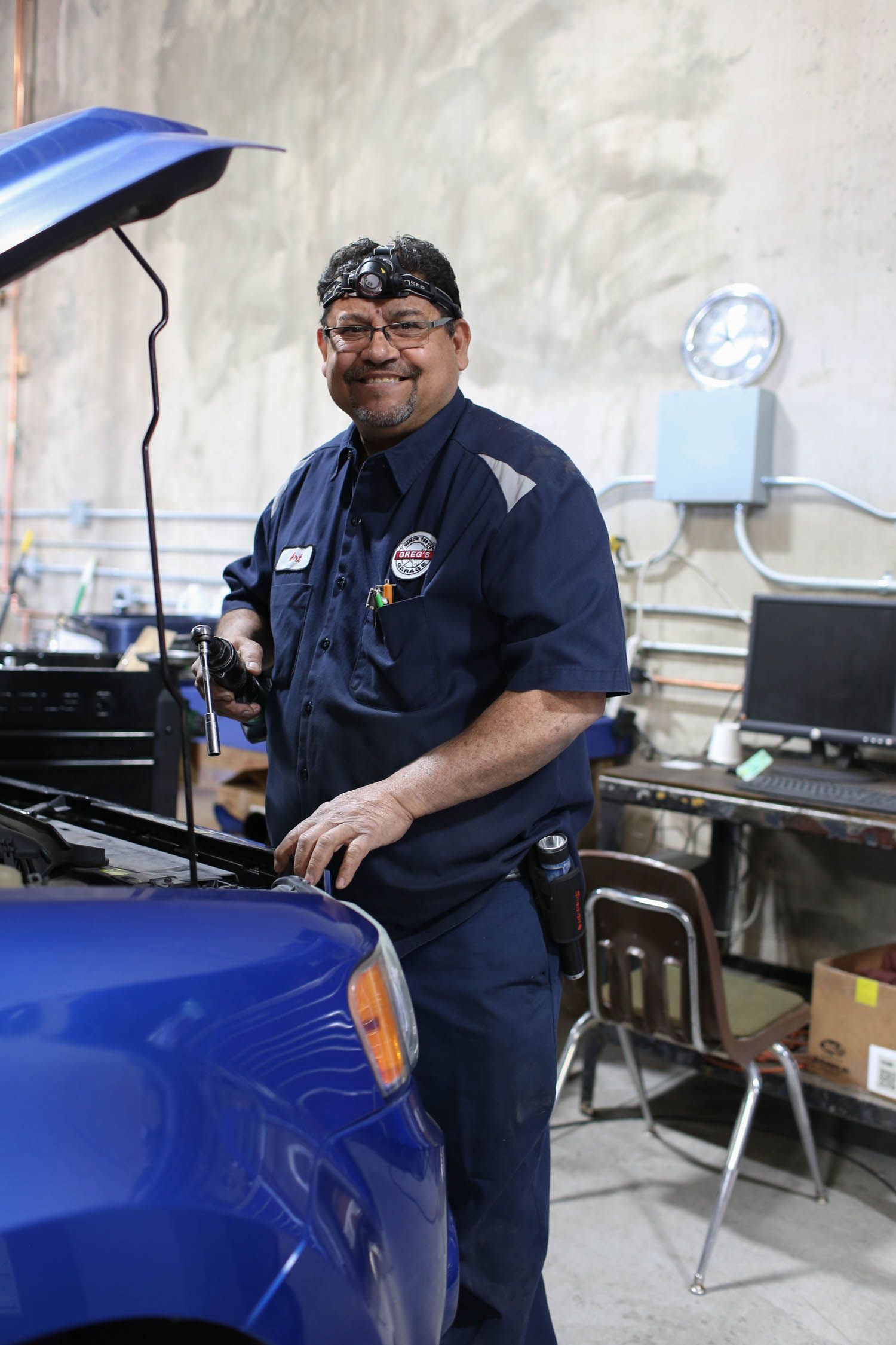 Auto mechanic in blue uniform smiles, leaning over a blue car in a garage. | Greg's Garage