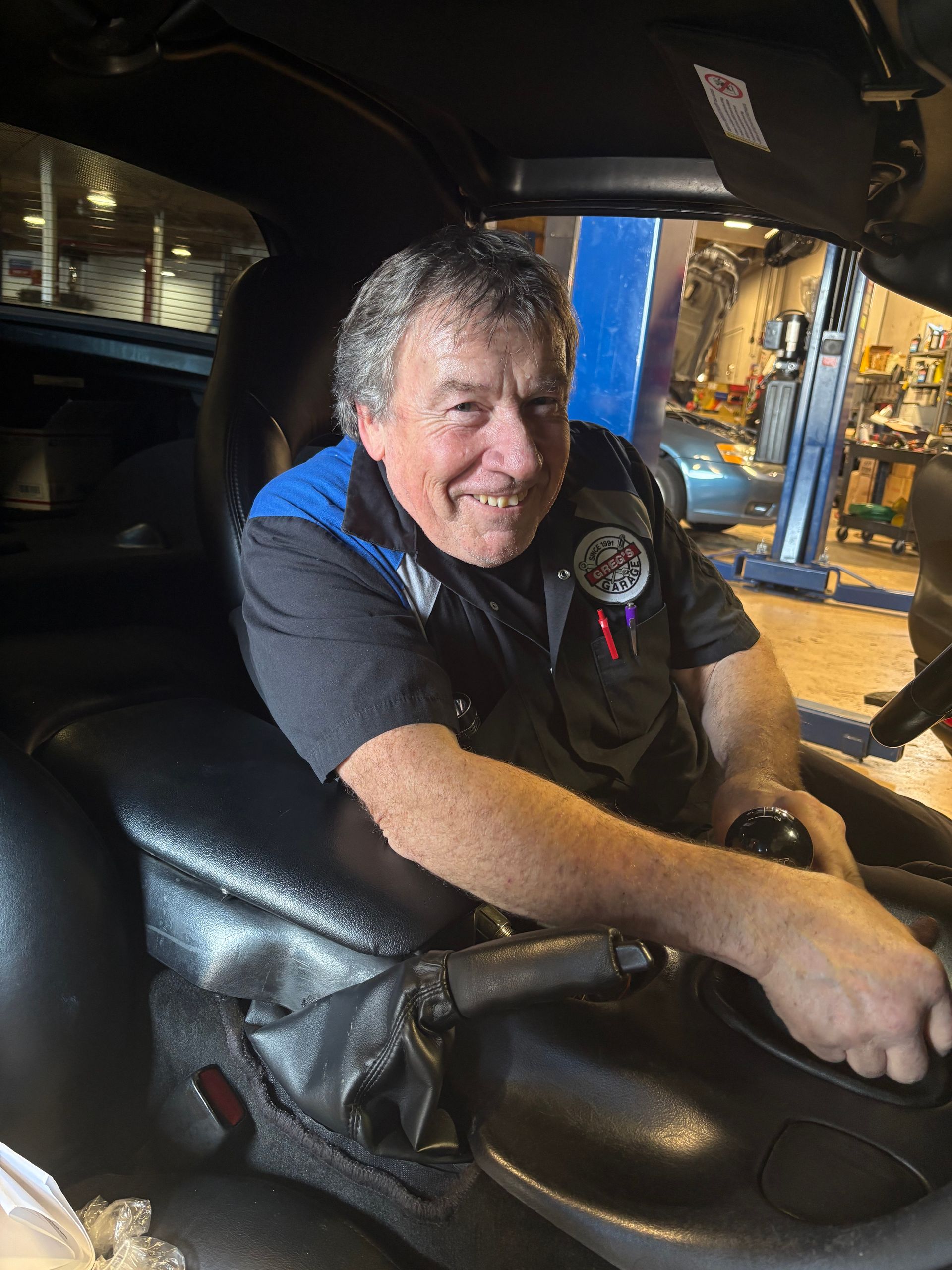 Mechanic sitting inside car, smiling. Wearing black and blue uniform, in a shop. | Greg's Garage