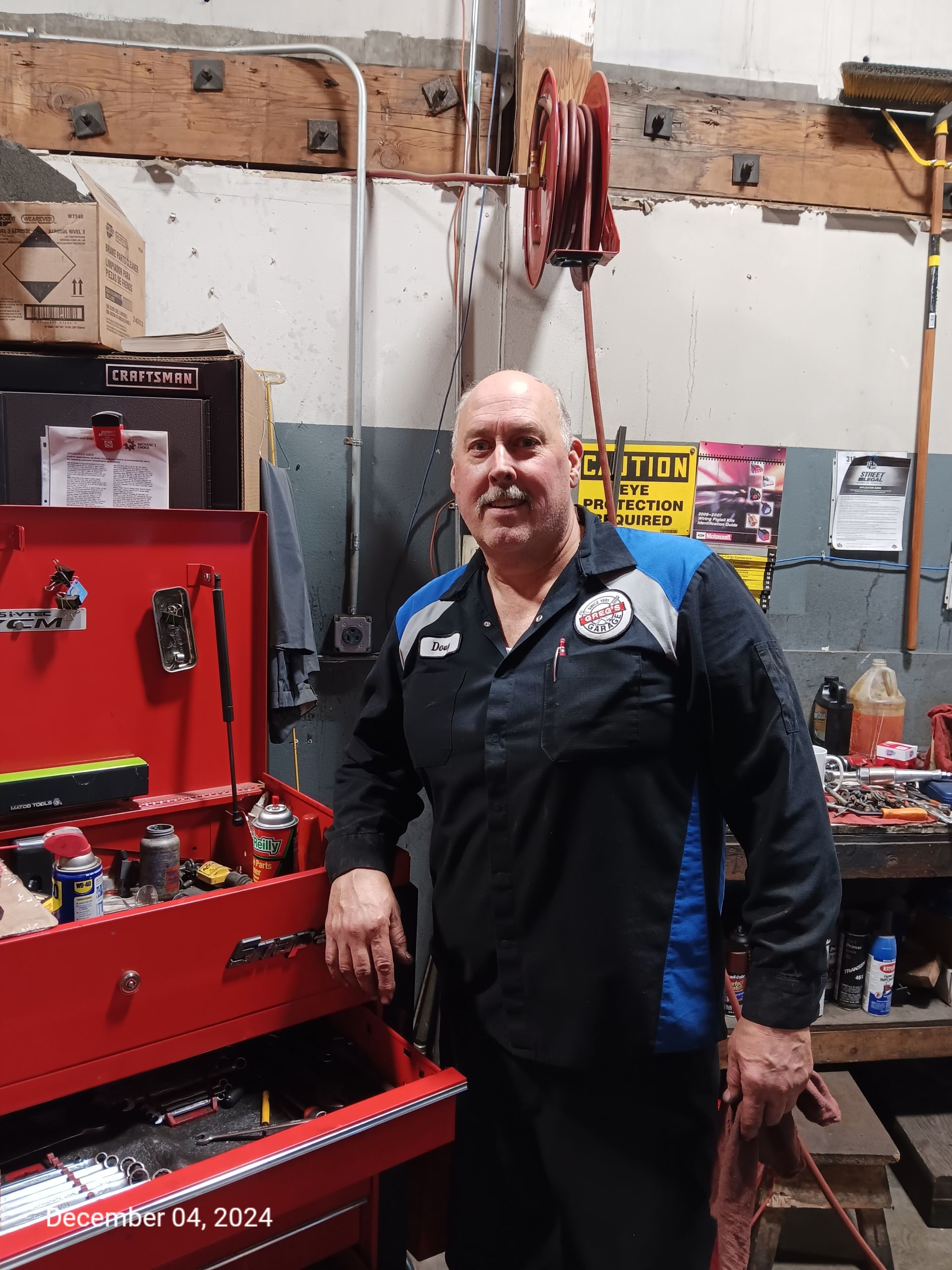 Doug Moss - A mechanic in a shop stands by a red toolbox, wearing a blue-accented uniform, smiling. | Greg's Garage