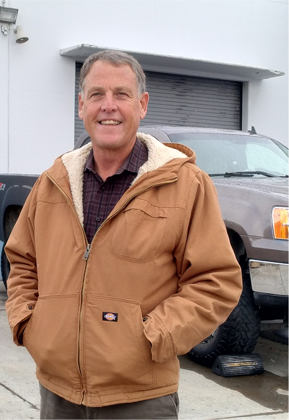 Man in brown coat smiling, standing in front of a building and a truck. | Greg's Garage