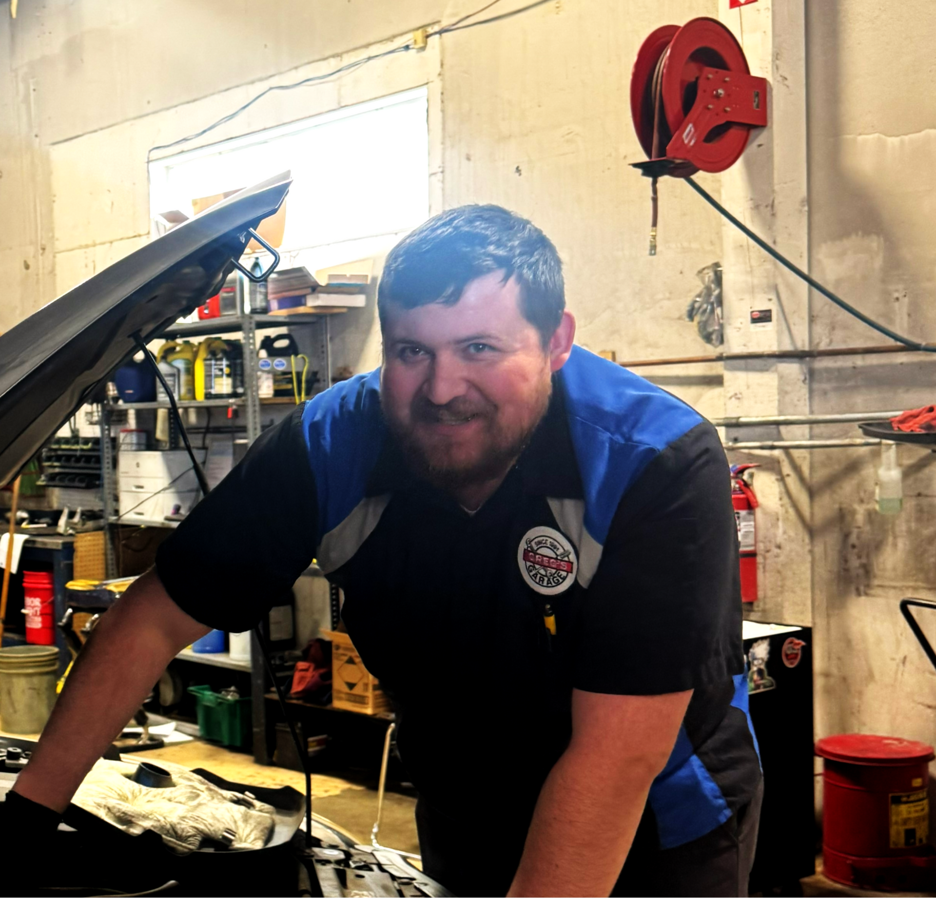 Mechanic smiles while working on a car in a garage; wearing blue-black uniform, standing next to open hood. | Greg's Garage