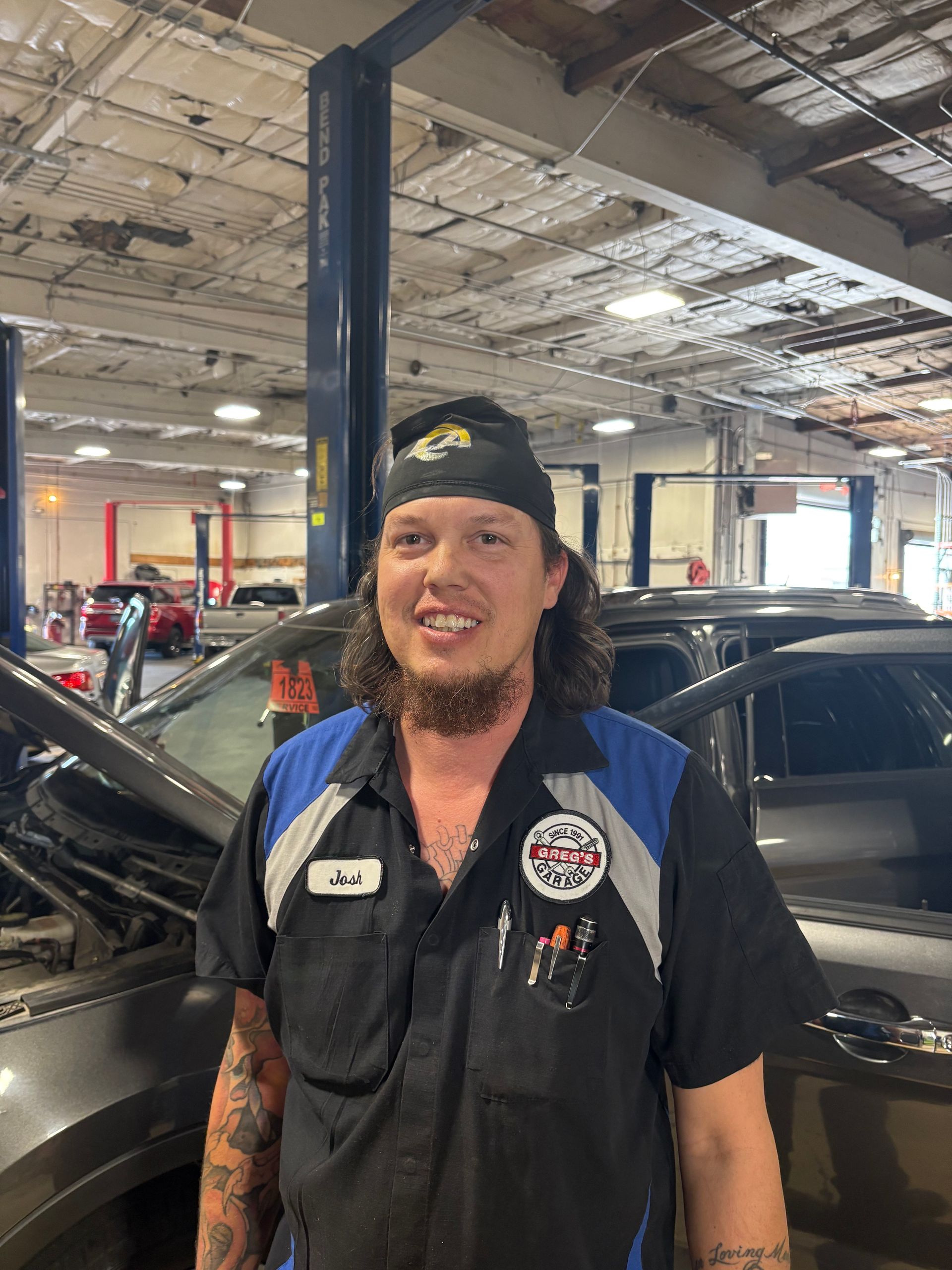 Mechanic in a shop smiling; wearing a beanie and uniform, standing near a car. | Greg's Garage