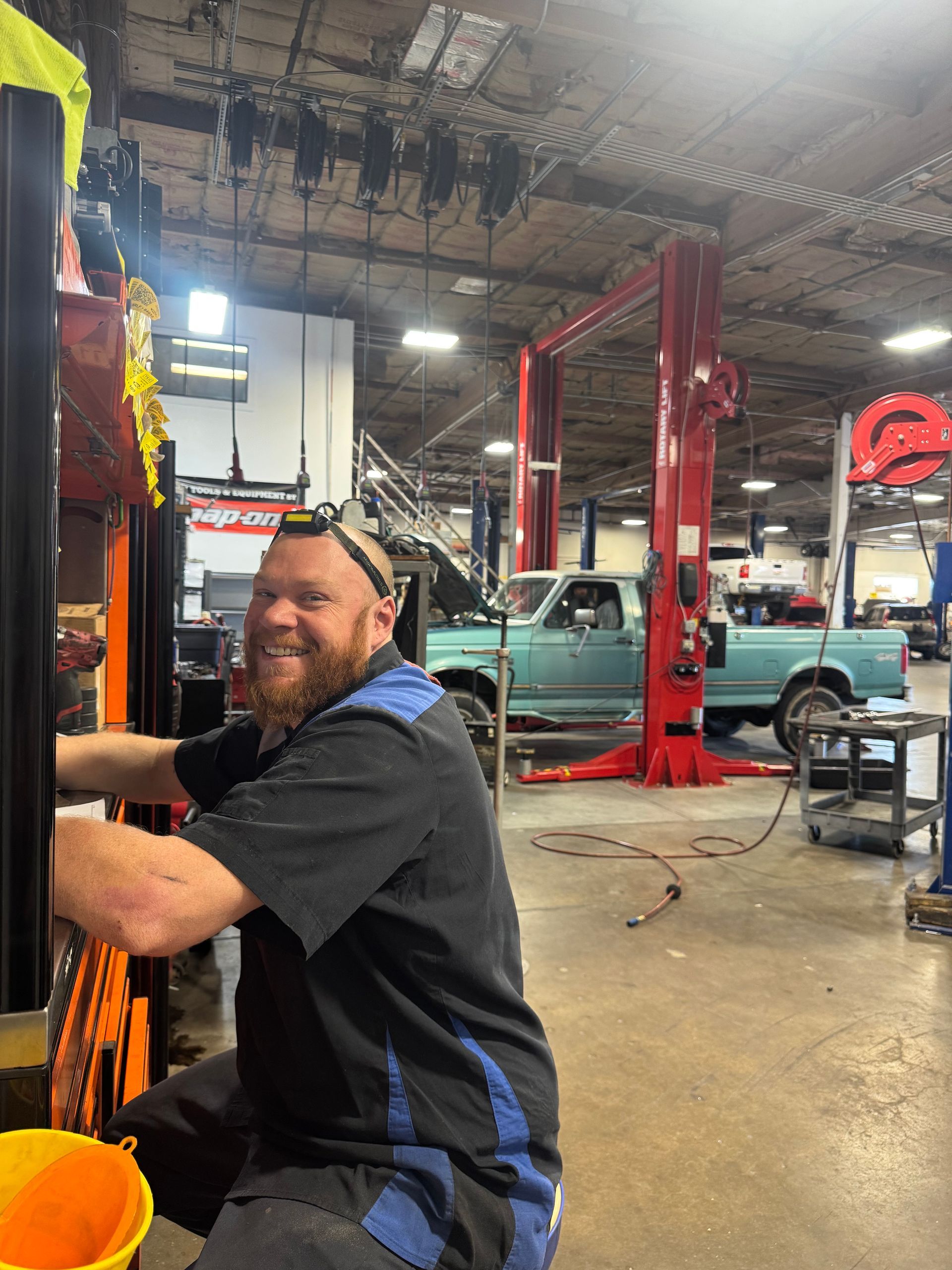 Michael Shea - Mechanic smiles while working on a lift in a garage with a light-blue pickup truck in the background. |Greg's Garage