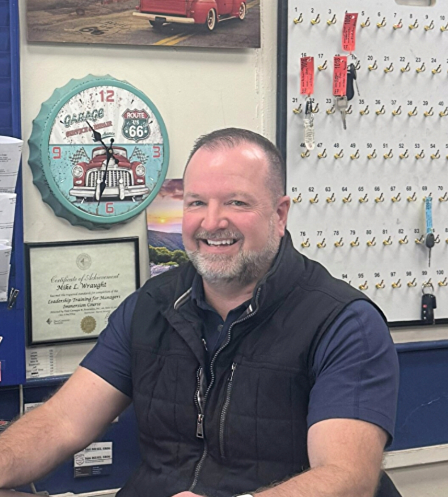 Mike Wraught - Man smiling, wearing vest, near a key rack and Route 66 clock.